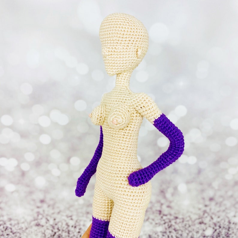 May include: A crocheted doll with a nude body and purple arms. The doll is made of yarn and has a simple design.