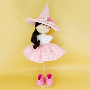 May include: A crocheted doll wearing a pink dress, a pink witch hat with flower accents, and pink shoes. The doll has brown hair and a white shirt.