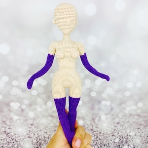 May include: A crocheted doll with a nude body and purple gloves and stockings. The doll is standing on a hand.