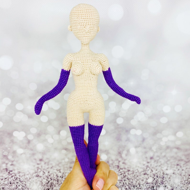 May include: A crocheted doll with a nude body and purple gloves and stockings. The doll is standing on a hand.