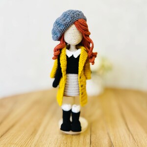 May include: A crocheted doll wearing a yellow cardigan, a black and white top, a gray skirt, a blue beret, and black boots. The doll has red hair and is standing on a wooden base.