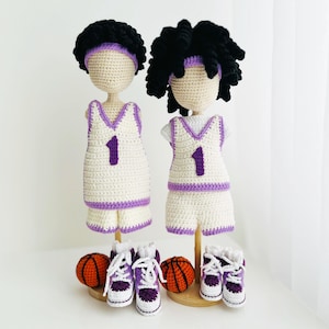 Peut inclure: Deux poupées au crochet vêtues d'uniformes de basket-ball blancs et violets, chacune avec le numéro 1. Elles ont des cheveux en fil noir et des bandeaux violets. Des ballons de basket miniatures et des baskets violets et blancs sont exposés en dessous.