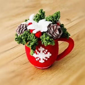May include: A red crocheted mug with a white snowflake design, filled with crocheted pine cones, holly, and white flowers.