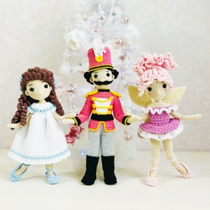 May include: Three crocheted dolls, a girl in a white dress, a girl in a pink dress, and a boy in a red and gold uniform. The dolls are standing on a white surface.