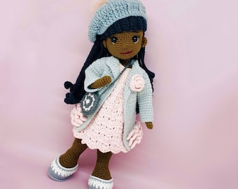 May include: Crocheted doll with dark skin, wearing a blue beret with a pink pom-pom, a light blue coat, and a pink dress. The doll has long black hair and is carrying a small purse. The doll is wearing white and gray shoes.