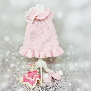 May include: A pink crocheted baby dress with a white crocheted collar and a matching headband. The dress is displayed on a white stand with a pink crocheted bunny and a pair of white crocheted booties.