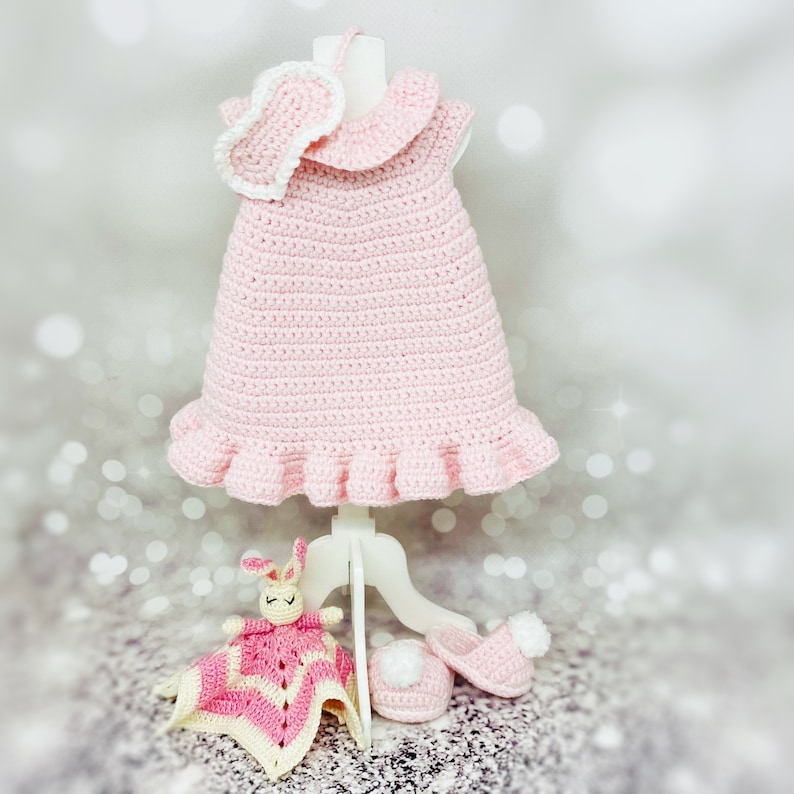 May include: A pink crocheted baby dress with a white crocheted collar and a matching headband. The dress is displayed on a white stand with a pink crocheted bunny and a pair of white crocheted booties.