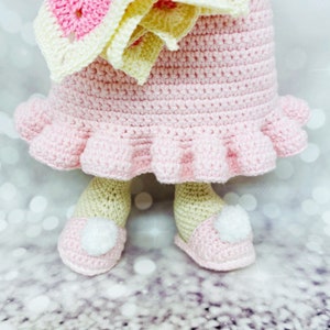 May include: A crocheted doll wearing a pink dress with a ruffled hem and a yellow and pink flower crown. The doll is wearing pink shoes with white pom-poms.