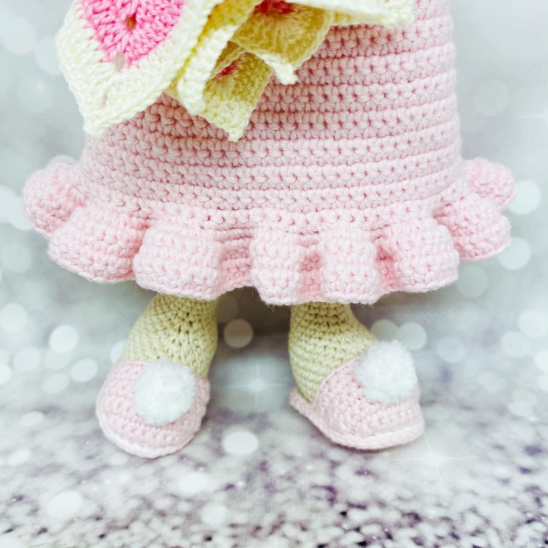 May include: A crocheted doll wearing a pink dress with a ruffled hem and a yellow and pink flower crown. The doll is wearing pink shoes with white pom-poms.