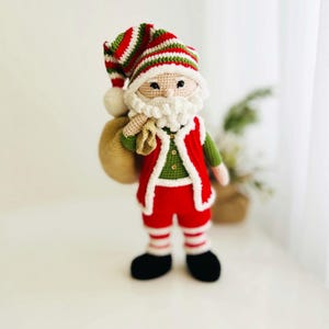 May include: A handmade crochet Santa Claus doll. The doll is wearing a red and white striped hat, a red vest with white trim, and red pants with white stripes. It has a white beard and a beige sack over its shoulder. The doll is standing upright.