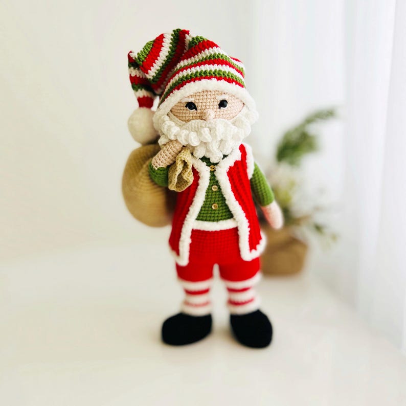 May include: A handmade crochet Santa Claus doll. The doll is wearing a red and white striped hat, a red vest with white trim, and red pants with white stripes. It has a white beard and a beige sack over its shoulder. The doll is standing upright.