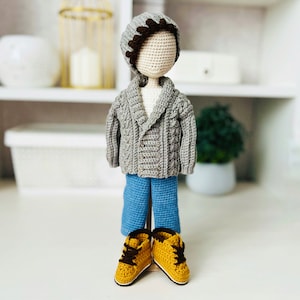 Può includere: Una bambola all'uncinetto che indossa un cardigan grigio, jeans blu e scarpe marroni. La bambola ha un cappello grigio con un bordo marrone.