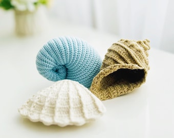 May include: Three crocheted seashell decorations. A light blue spiral shell, a beige conch shell, and a white scallop shell. The shells are handmade and have a textured appearance.