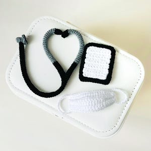 Peut inclure: Ensemble médical crocheté comprenant un stéthoscope, une tablette et un masque. Le stéthoscope est noir et gris, en forme de cœur. La tablette est blanche avec un bord noir, et le masque est blanc. Le tout est posé sur une surface blanche.