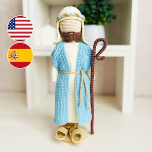 May include: Crocheted amigurumi shepherd doll with a blue and white robe, a brown staff, and a brown beard. The doll is standing on a wooden base.