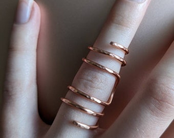 Op de afbeelding: Een gehammerde koperen ring met een spiraalvormig ontwerp. De ring wordt aan een vinger gedragen.