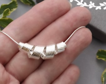 May include: A silver necklace with a unique design. The necklace features a series of textured, curled silver elements strung on a delicate chain. The box in the background reads "MADE BY FEE SILVER JEWELLERY".