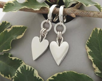 May include: A pair of silver heart-shaped earrings. Each earring has a polished heart pendant hanging from a hoop and a small ring. The earrings are set against a grey background with green and white leaves.