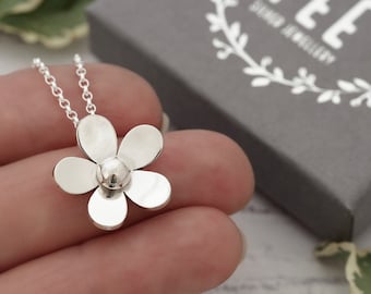 May include: A silver flower pendant necklace. The necklace features a five-petal flower design with a small, round centre. The chain is delicate and silver. The background includes a grey box with the text "FEE SILVER JEWELLERY".