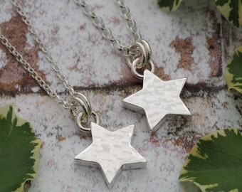 May include: Two silver star pendant necklaces. Each necklace features a hammered star pendant suspended from a delicate chain. The pendants are silver and have a textured surface. The necklaces are displayed on a neutral background with green leaves.