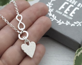 May include: A silver necklace featuring an infinity symbol and a hammered heart pendant. The necklace is on a chain and is displayed with a gray box that has the text "MADE BY FEE SILVER JEWELLERY".
