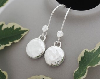 May include: A pair of silver drop earrings. Each earring has a hammered, circular pendant hanging from a curved hook. The earrings are set against a grey background with green and white leaves.