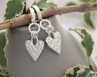 May include: Silver heart earrings with a textured finish. The earrings are suspended from silver hoops and are displayed on a grey vase, with green and white foliage in the background.