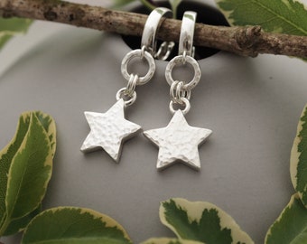 May include: Silver star earrings with a hammered finish, suspended from a hoop and a small ring. The background is a soft grey, with green leaves and a branch. A stylish accessory.
