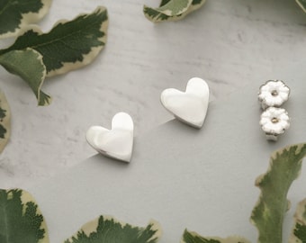 May include: A pair of polished silver heart-shaped stud earrings. The earrings are displayed with two matching earring backs. Green and white leaves are scattered around the earrings on a light gray surface.