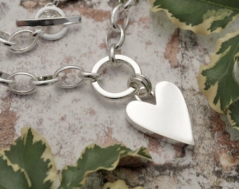 May include: A silver-toned necklace with a heart pendant. The necklace features a toggle clasp and a chain with oval links. The heart pendant is smooth and polished. The background includes green and white leaves.