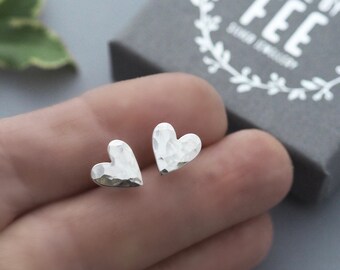 May include: A pair of silver heart-shaped stud earrings with a hammered texture. The earrings are displayed on a person's hand, with a gray box in the background that reads "MADE BY FEE SILVER JEWELLERY".