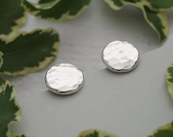 May include: Two silver stud earrings with a hammered texture. The earrings are circular with a slightly uneven surface. The background features green and white variegated leaves, creating a natural, elegant display. The earrings are presented on a light grey surface.