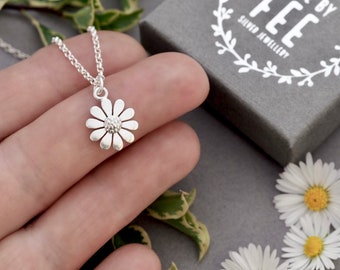 May include: A silver daisy pendant necklace. The daisy pendant has a detailed center and petals. The necklace is displayed on a gray surface with a gray box that reads "MADE BY FEE SILVER JEWELLERY".