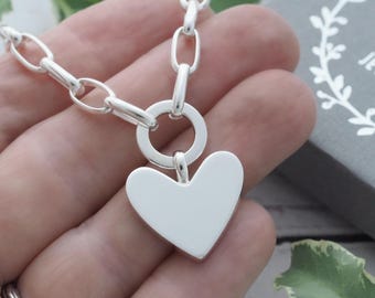 May include: A silver heart pendant necklace with a large heart-shaped charm. The necklace has a chunky chain design and a circular clasp. The heart charm is smooth and polished. The jewelry is displayed against a neutral background.