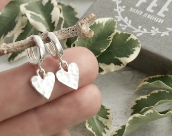 May include: A pair of silver hoop earrings with hammered heart pendants. The earrings are displayed on a small branch, with green and white leaves in the background. The earrings are a simple, elegant design.