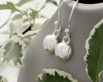 May include: A pair of silver drop earrings with a round, textured design. The earrings hang from curved hooks and are displayed against a gray background with green and white foliage.