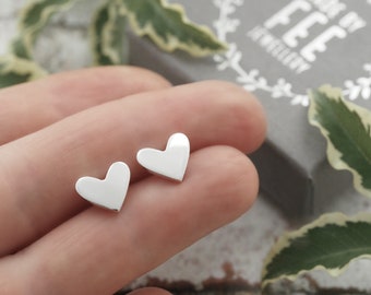May include: A pair of small, shiny, silver heart-shaped stud earrings. The earrings are displayed on a person's fingers, with a gray box in the background. The box has the text "MADE BY FEE JEWELLERY".