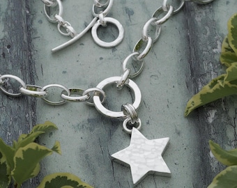 May include: A silver-toned necklace with a hammered star pendant. The necklace features a toggle clasp and a series of oval-shaped links. The star pendant is textured and hangs from a large ring. The jewelry is displayed on a gray and green surface.