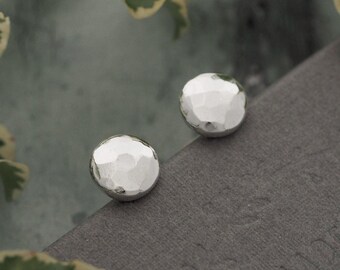 May include: Two silver stud earrings with a hammered texture. The earrings are round and have a faceted surface, reflecting light. The earrings are displayed on a gray surface, with green and white foliage in the background.