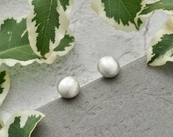 May include: Two small, round, silver-colored stud earrings. The earrings are on a gray surface, with green and white leaves in the background. The earrings have a smooth, matte finish.