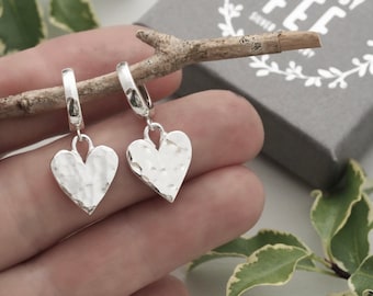 May include: A pair of silver heart-shaped earrings with a hammered texture. The earrings are suspended from small hoops. The background includes a small branch, a gray box, and green foliage.