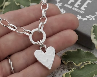 May include: A silver necklace with a hammered heart pendant. The necklace features a chunky chain and a large circular clasp. The heart pendant has a textured surface. The image also shows a ring and a box that says "MADE BY FEE SILVER JEWELLERY."