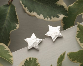 May include: A pair of silver star-shaped stud earrings with a hammered texture. The earrings are displayed on a white surface, surrounded by green and white variegated leaves. The earrings are a celestial-themed accessory.