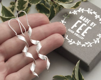 May include: A pair of silver, spiral-shaped earrings with textured surfaces. The earrings are displayed in a hand, with a gray box that reads "MADE BY FEE SILVER JEWELLERY" in the background. Green and white leaves are in the background.