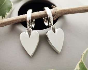 May include: Silver heart-shaped drop earrings. The earrings hang from small hoops and are presented on a twig against a grey backdrop. The heart pendants have a smooth, polished surface. The earrings are a romantic accessory.