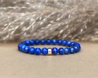 May include: A stretch bracelet composed of round, deep blue lapis lazuli beads with visible natural flecks, featuring a single smooth, polished gold-tone cylindrical spacer bead centered at the front of the circular arrangement.