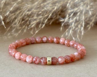May include: A beaded bracelet composed of small, round, peach-colored sunstone beads with a single, smooth, cylindrical gold-toned metal spacer bead positioned near the center of the circular strand.