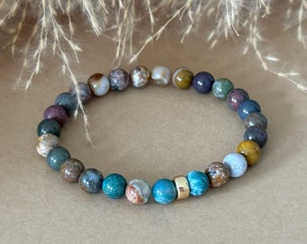May include: A beaded bracelet featuring a variety of round, multi-colored natural stone beads in shades of blue, green, brown, and purple. The bracelet includes a single smooth, cylindrical gold-toned metal bead accent positioned near the center.