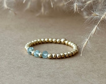 May include: A gold beaded ring with three light blue gemstones. The ring is set against a neutral background with dried ornamental grass in the upper portion of the image. The ring is a delicate piece of jewelry.