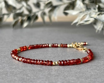 May include: A delicate bracelet featuring small, faceted red beads interspersed with gold-colored beads. The bracelet is secured with a gold-colored clasp. The jewelry is displayed on a gray surface with a blurred background of greenery.